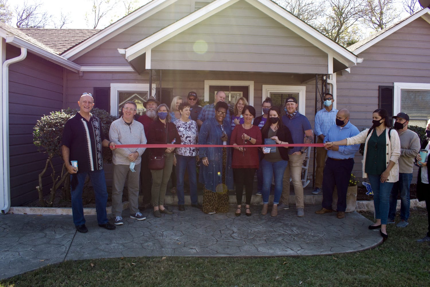 Various individuals standing in front of a house aka Amira's House, female holding scissors to cut the ribbon for grande opening.